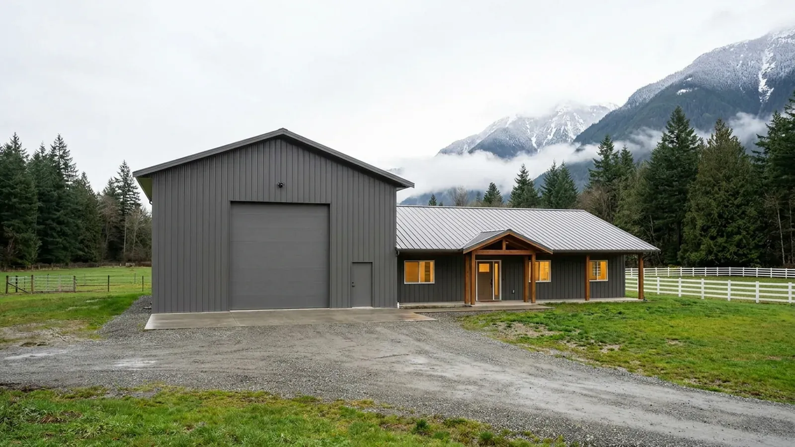 A barndominium with a tall, deep RV garage bay and an oversized overhead door beside a single-storey living wing, forested mountains behind.